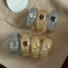Six textured bangle-style watches arranged on a beige fabric surface, with two silver-toned and two gold-toned bracelets plus two additional silver and gold bracelets showing different dial colors. A white circular card with the brand name Reminisce is partially visible in the top left.