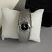 A silver-toned women's watch with a textured, hammered metal band and a black dial, displayed on a white textured cushion.