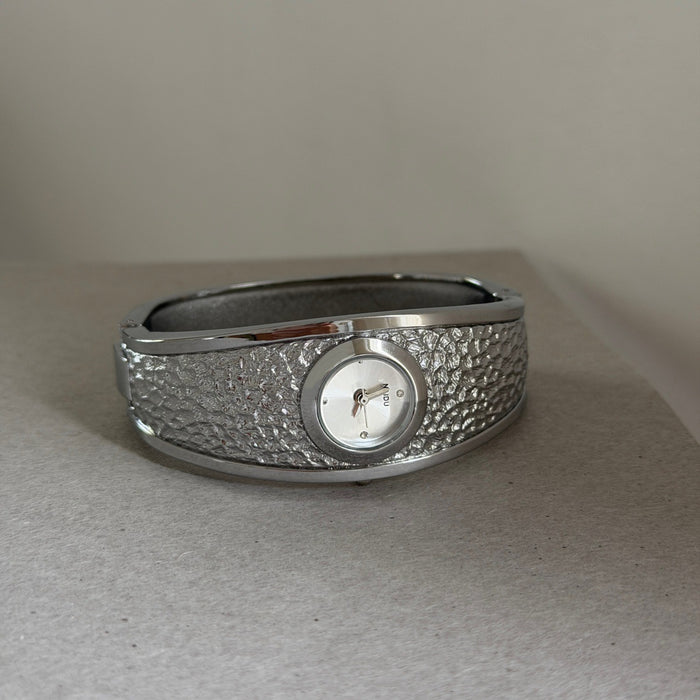 A silver-toned wide cuff bracelet watch with a textured, hammered outer band, a polished inner lining, and a small round white dial face.