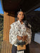 A woman stands on a wooden deck wearing a white long-sleeve blouse with large black polka dots and holding a small woven handbag, with a modern wooden building and hills in the background.