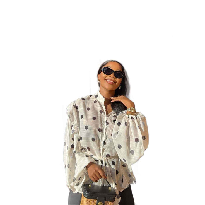 A woman wearing a white long-sleeve blouse with black polka dots, black sunglasses, and a black handbag, set against a plain white background.