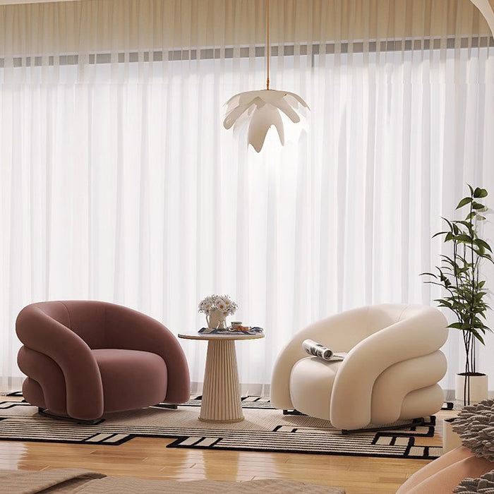 A modern living room scene with two sculptural armchairs, one dusty rose and one cream, arranged around a small round side table on a patterned rug. The room has sheer white curtains, a white leaf-shaped pendant light, and a potted plant.