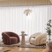A modern living room scene with two sculptural armchairs, one dusty rose and one cream, arranged around a small round side table on a patterned rug. The room has sheer white curtains, a white leaf-shaped pendant light, and a potted plant.