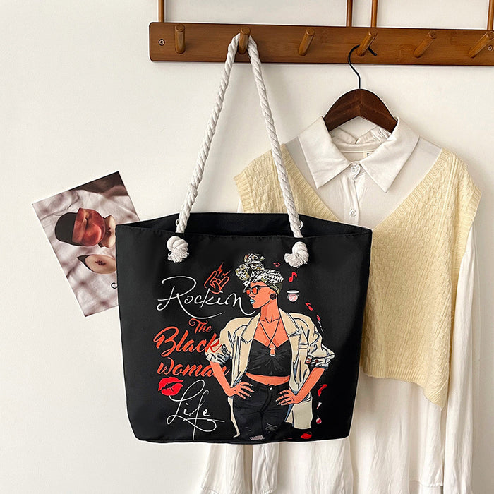 Black tote bag with a graphic design of a woman and text, hanging on a wooden coat rack with a shirt and photo in the background.