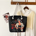 Black tote bag with a graphic design of a woman and text, hanging on a wooden coat rack with a shirt and photo in the background.