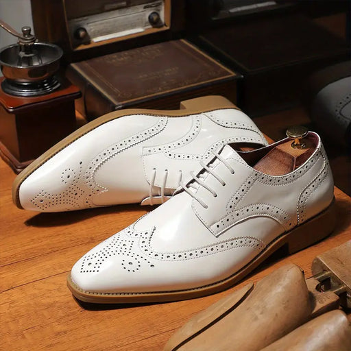 A pair of white leather wingtip oxford shoes with decorative brogue perforations, displayed on a wooden surface with vintage props in the background.