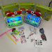 A children's tablet kit displayed on a white surface with its retail boxes. The kit includes a tablet in a green and white protective case, a tablet in a pink and white protective case, two colorful toy swords, a green wrist strap, sticker sheets, a power adapter, a charging cable, and sunglasses.