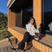 A woman sits on the wooden deck of a modern cabin wearing a white ruffled blouse with black polka dots, black wide-leg trousers, and black pointed heels, holding a woven clutch.