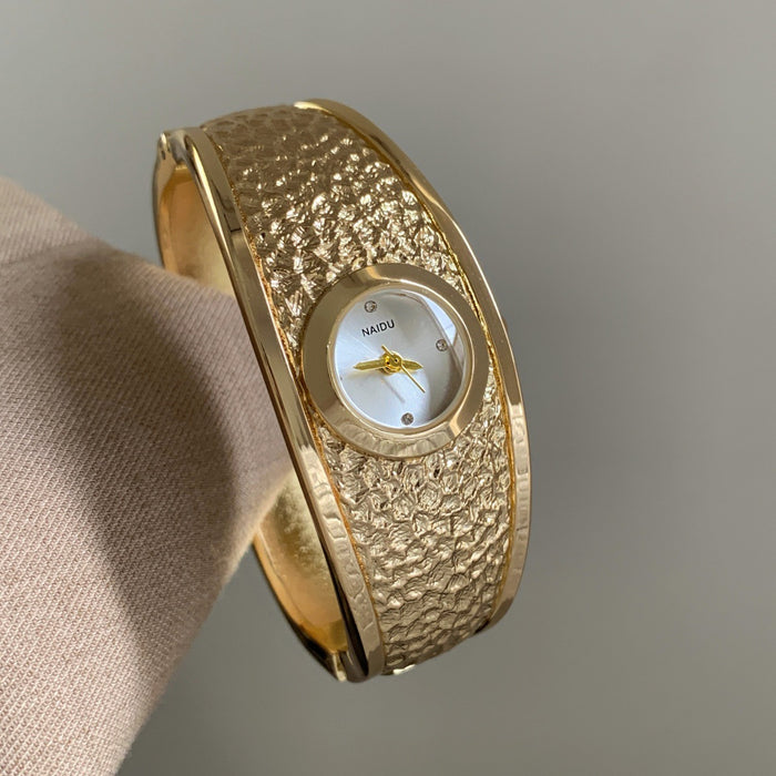 A gold-toned textured bangle watch with a white dial and gold hands, shown resting on a beige fabric surface against a neutral gray background.