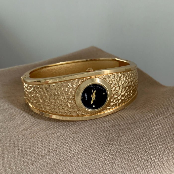 A gold-toned textured bangle watch with a black dial, resting on a beige fabric surface against a neutral gray background.