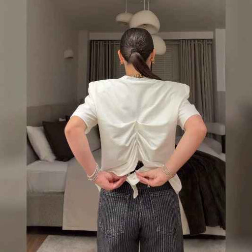 A woman viewed from behind wearing a white short-sleeve top with a ruched back and tie detail, paired with dark pinstriped pants in a modern bedroom setting.