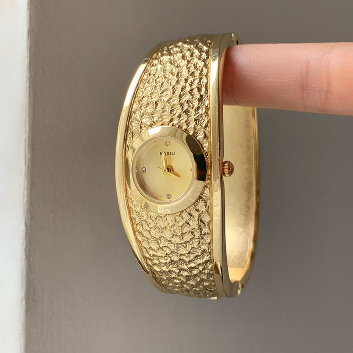 A gold-plated bangle watch with a textured, hammered outer band and a smooth dial, held by a person's finger against a neutral gray background.