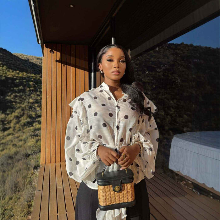 A woman stands on a wooden deck outdoors wearing a white long-sleeve blouse with black polka dots, holding a small woven handbag. Mountains and a modern building are visible in the background.