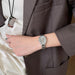 A silver-toned analog watch with a matching metal link bracelet worn on a person's wrist, shown against a brown blazer and cream satin blouse.