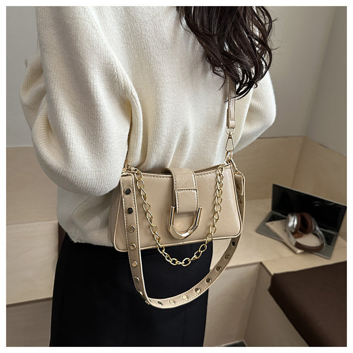 A person wearing a cream knit sweater and black skirt carries a beige shoulder bag with a gold chain strap and studded details. The photo is taken indoors with a light wood surface and a small mirror in the background.