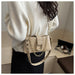 A person wearing a cream knit sweater and black skirt carries a beige shoulder bag with a gold chain strap and studded details. The photo is taken indoors with a light wood surface and a small mirror in the background.