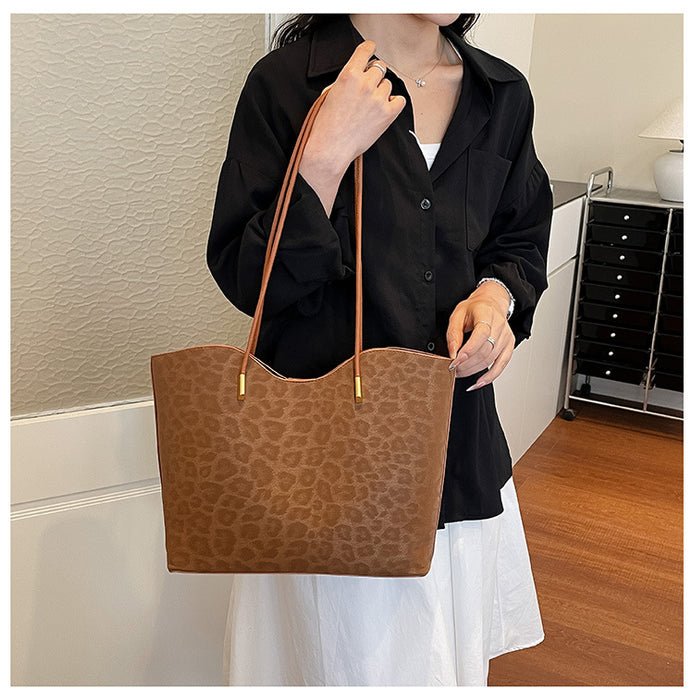 A person wearing a black button-up shirt and white skirt holds a large brown leopard-print tote bag with tan shoulder straps and gold-tone hardware in an indoor room setting.