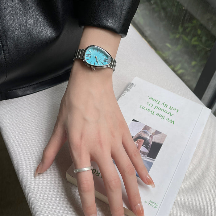 A silver-tone metal watch with a bright turquoise blue dial is worn on a person's wrist, resting on a white textured surface next to an open book.