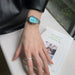 A silver-tone metal watch with a bright turquoise blue dial is worn on a person's wrist, resting on a white textured surface next to an open book.