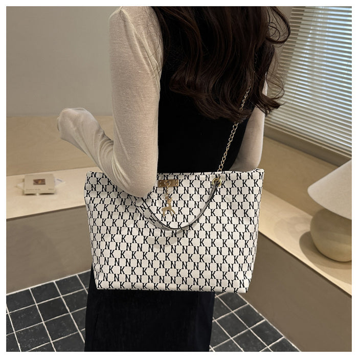 A woman wearing a cream long-sleeve top and black dress carries a white tote bag with a black geometric pattern and a gold chain strap in an indoor setting.