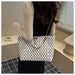 A woman wearing a cream long-sleeve top and black dress carries a white tote bag with a black geometric pattern and a gold chain strap in an indoor setting.