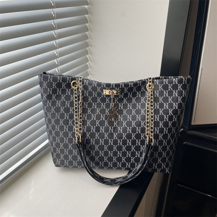 A structured tote bag with a dark gray and black monogram pattern, featuring gold-tone chain straps and a central logo plaque, resting on a windowsill next to window blinds.