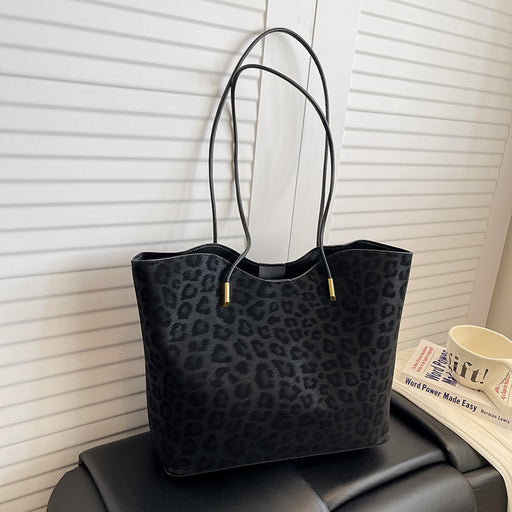 A black leopard-print tote bag with thin shoulder straps and small gold hardware, resting on a black leather chair in front of white horizontal blinds. A book and a white mug are partially visible on the right side.