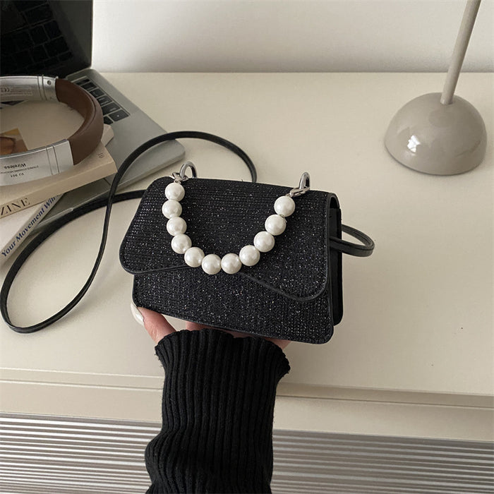 A black glittery mini handbag with a white faux-pearl beaded collar and strap, held by a hand wearing a black ribbed sweater on a light beige desk with a laptop and lamp in the background.