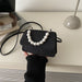 A black glittery mini handbag with a white faux-pearl beaded collar and strap, held by a hand wearing a black ribbed sweater on a light beige desk with a laptop and lamp in the background.