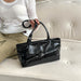 A black glossy leather tote bag with double top handles and a front zippered pocket, held by a person sitting on a cream tufted sofa.