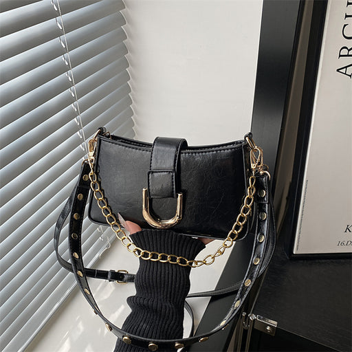 A black faux-leather shoulder bag with a gold-tone chain strap and studded details, held by a hand wearing a black ribbed sweater in front of window blinds.