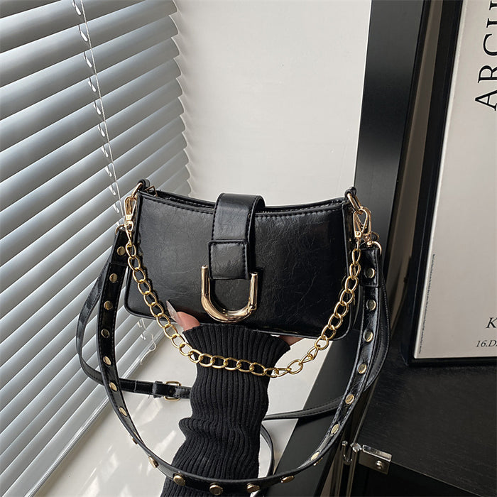 A black faux-leather shoulder bag with a gold-tone chain strap and studded details, held by a hand wearing a black ribbed sweater in front of window blinds.