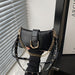 A black faux-leather shoulder bag with a gold-tone chain strap and studded details, held by a hand wearing a black ribbed sweater in front of window blinds.