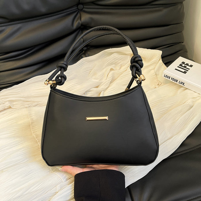 A black leather handbag with knotted top handles and a small gold bar detail on the front, held by a hand over a cream-colored textured fabric with a black leather sofa in the background.
