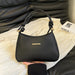 A black leather handbag with knotted top handles and a small gold bar detail on the front, held by a hand over a cream-colored textured fabric with a black leather sofa in the background.