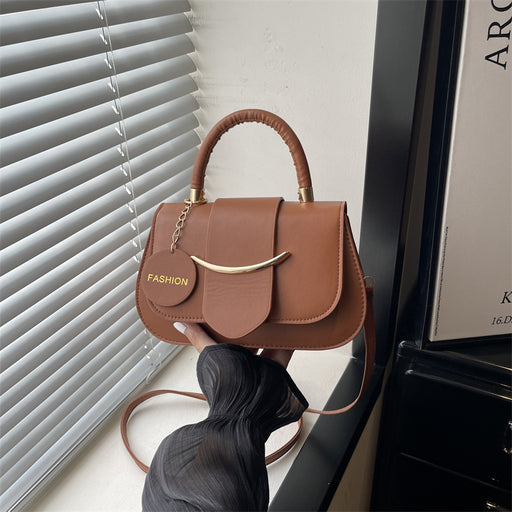 A brown leather handbag with a curved top handle, gold-tone hardware, and a small round charm reading 'FASHION', held by a person wearing a dark sleeveless top near a window with blinds.