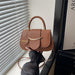 A brown leather handbag with a curved top handle, gold-tone hardware, and a small round charm reading 'FASHION', held by a person wearing a dark sleeveless top near a window with blinds.