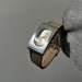 Vintage-style wristwatch with brown leather strap held by a gloved hand against a gray background