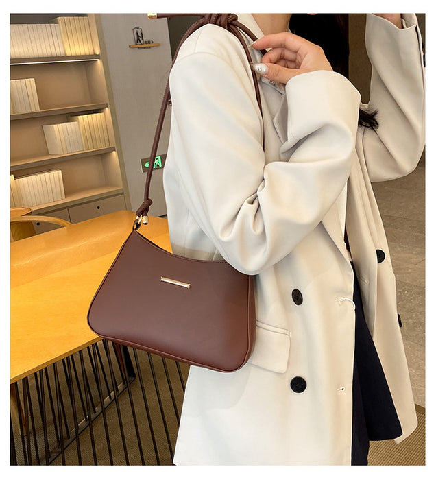 A person wearing a cream-colored double-breasted coat carries a brown shoulder bag with a small gold accent in a modern indoor setting.
