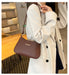 A person wearing a cream-colored double-breasted coat carries a brown shoulder bag with a small gold accent in a modern indoor setting.