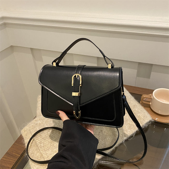 A black leather handbag with a top handle and shoulder strap, held by a hand. The bag features a front flap with a central gold-tone buckle closure and is styled on a textured beige surface with a white wall and wooden table in the background.