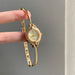 A gold-tone NAOJI watch with a curved bangle bracelet, held by a person's fingers against a neutral gray background.