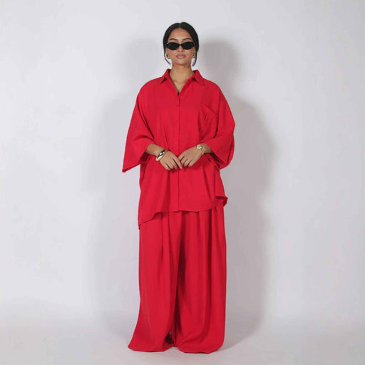 A model wearing a bright red two-piece matching set consisting of a long-sleeve button-up shirt and wide-leg trousers, styled with black sunglasses and gold jewelry against a plain light gray studio background.
