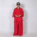 A model wearing a bright red two-piece matching set consisting of a long-sleeve button-up shirt and wide-leg trousers, styled with black sunglasses and gold jewelry against a plain light gray studio background.