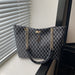 A structured tote bag with a dark gray and black monogram pattern, featuring gold-tone chain straps and a central logo plaque, resting on a windowsill next to window blinds.