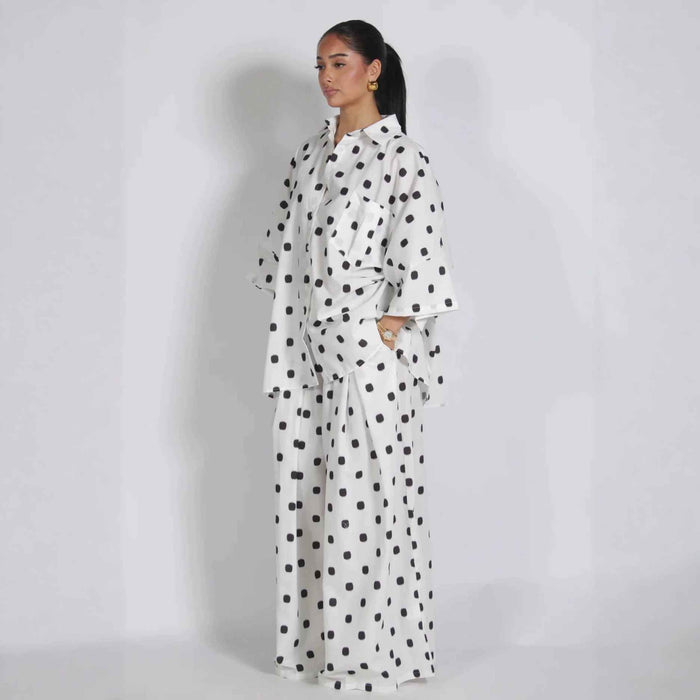 A woman modeling a white long-sleeve shirt and matching wide-leg trousers set with a black polka dot pattern, standing against a light gray studio background.