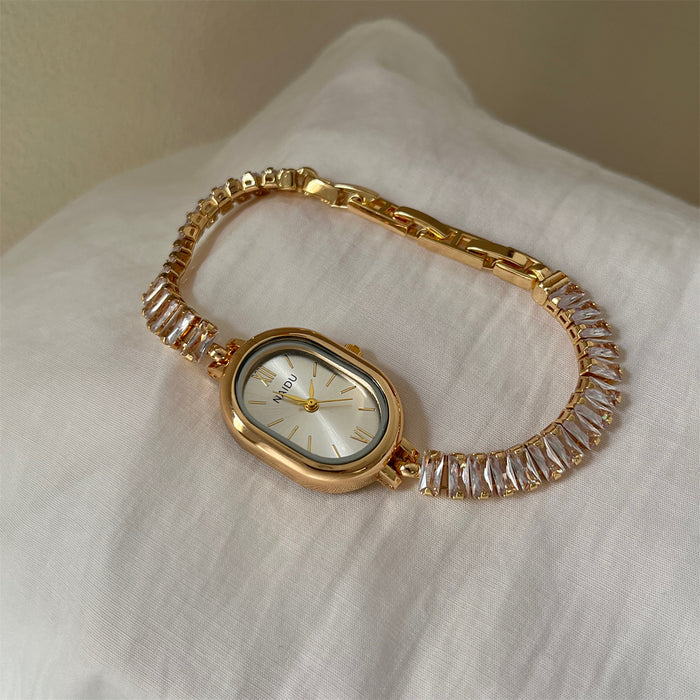 A gold-tone women's watch with a bezel-encased oval face and a link bracelet covered in clear rectangular crystals, resting on a white textured fabric surface.