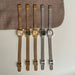 Four vintage-style wristwatches with mesh metal straps are arranged vertically on a light surface with a brown fabric prop behind them. The watches have gold-tone and silver-tone cases and straps, with one green dial and two white dials.