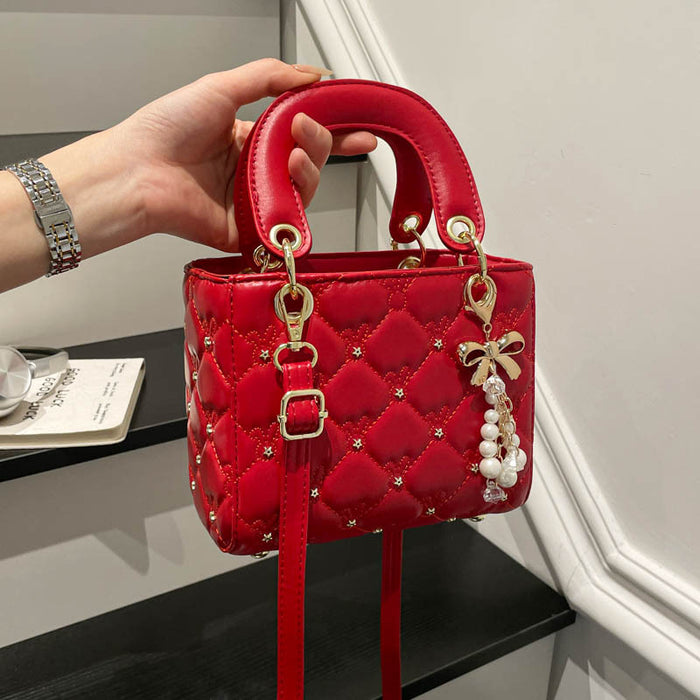 A red quilted top-handle handbag with gold-tone hardware, rhinestone accents, and a pearl bow charm, held by a hand in an indoor setting.