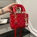 A red quilted top-handle handbag with gold-tone hardware, rhinestone accents, and a pearl bow charm, held by a hand in an indoor setting.